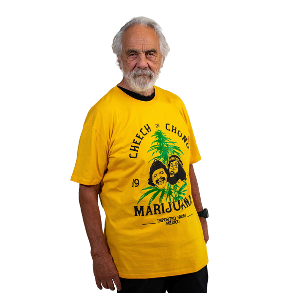 Tommy stand wearing a yellow shirt with black font that reads Cheech and Chong In the middle of the shirt is a drawn picture of Cheech and Chong's heads that sits in front of a tall green hemp plant.
