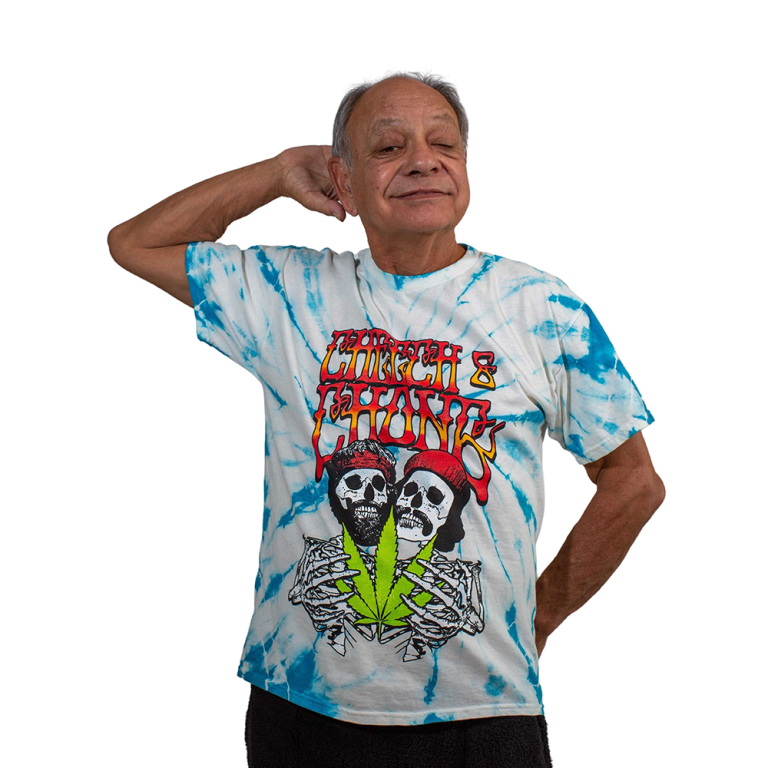 Cheech poses, wearing a blue and white tie died t-shirt with red and yellow writing that says Cheech and Chong and a skelton Cheech and Chong below that.