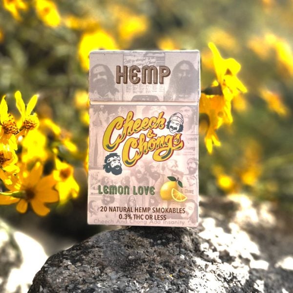 A pack of Granddaddy Grape Hemp Cigarettes with 'Lemon Love' flavor positioned on a rock, surrounded by blurred yellow flowers. The pack is labeled as containing natural hemp with 0