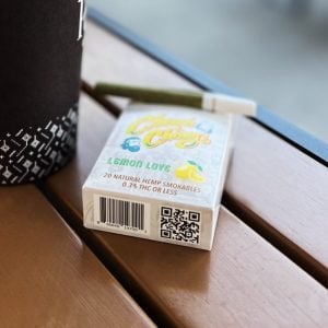A pack of Granddaddy Grape Hemp Cigarettes on a wooden surface next to a cup, with a green smoking device resting on top. A QR code is visible on the side of the package.