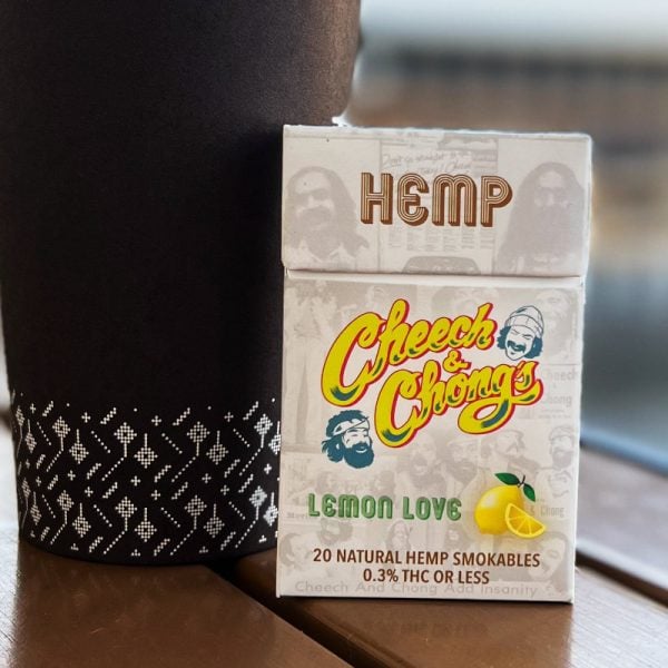 A pack of Granddaddy Grape hemp cigarettes on a wooden table, next to a black textured cup. The packaging features a yellow lemon graphic and notes of containing