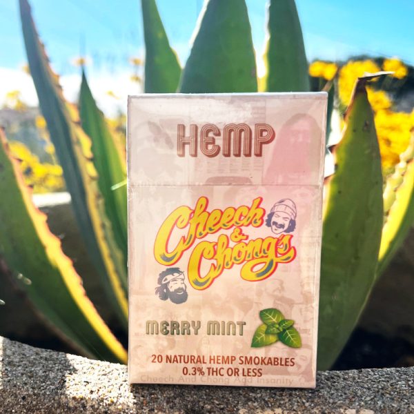 A package of "Granddaddy Grape Hemp Cigarettes" with a 0.3% THC content, placed in front of bright green aloe vera plants under a clear, sunny sky.