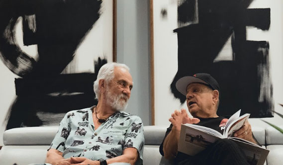 Tommy Chong and Ceech Marin, sit on a couch in front of a modern painting. Tommy smiles as Cheech holds a book while talking.
